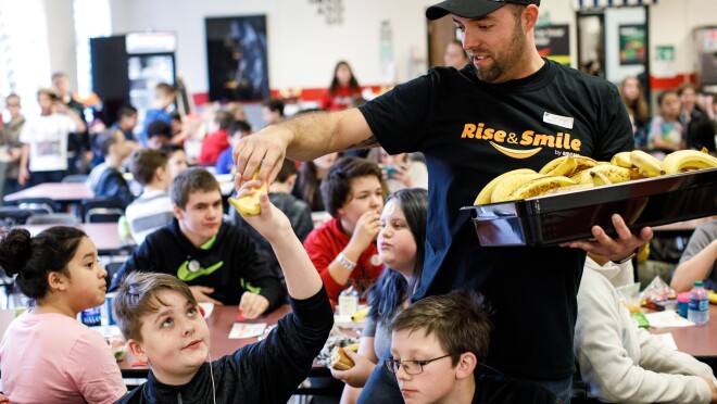 在一所学校,一名男子端着一盘香蕉,穿着Rise & Smile t恤,把香蕉递给一名学生