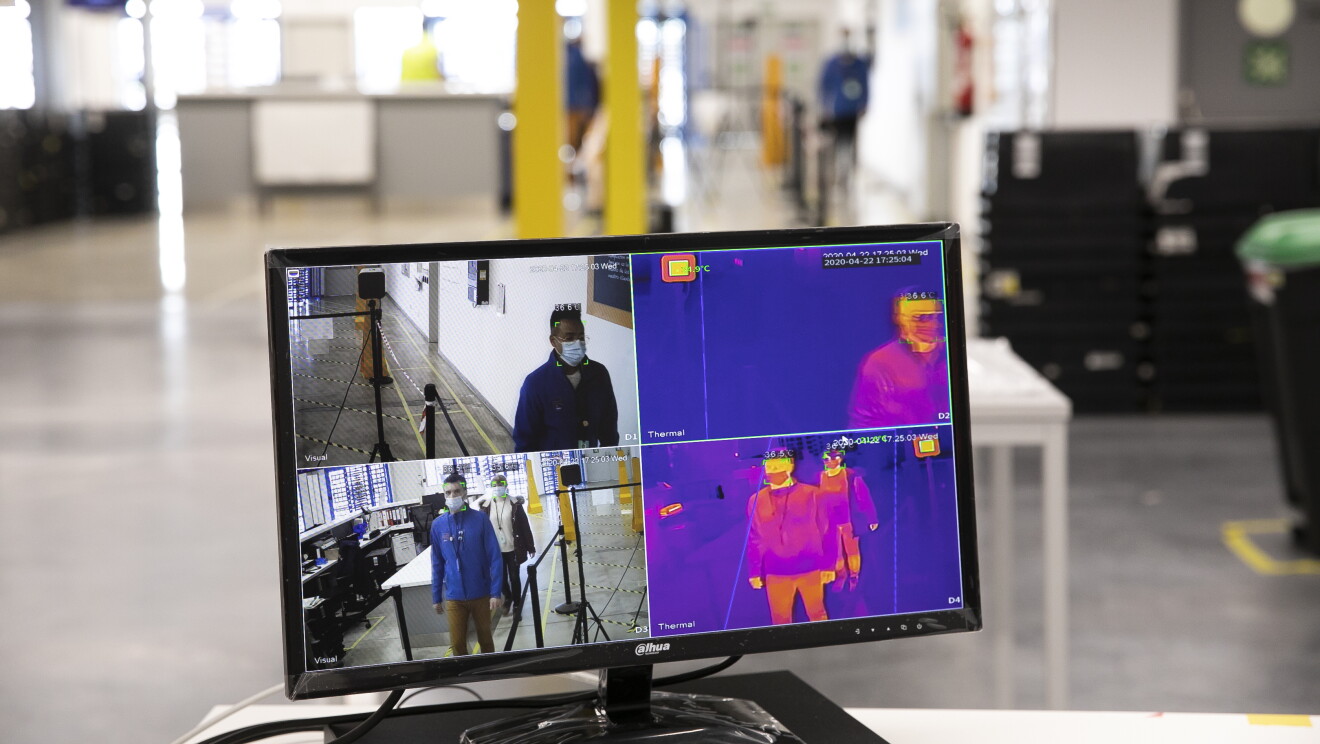 Hemos incluido controles de temperatura diarios a todos los trabajadores antes de acceder a los centros. En primer plano una pantalla de ordenador con cuatro imágenes, dos de las personas pasando un control y al lado las mismas imágenes pero observando el control de temperatura. De fondo desenfocado se ve la entrada a un centro logístico.