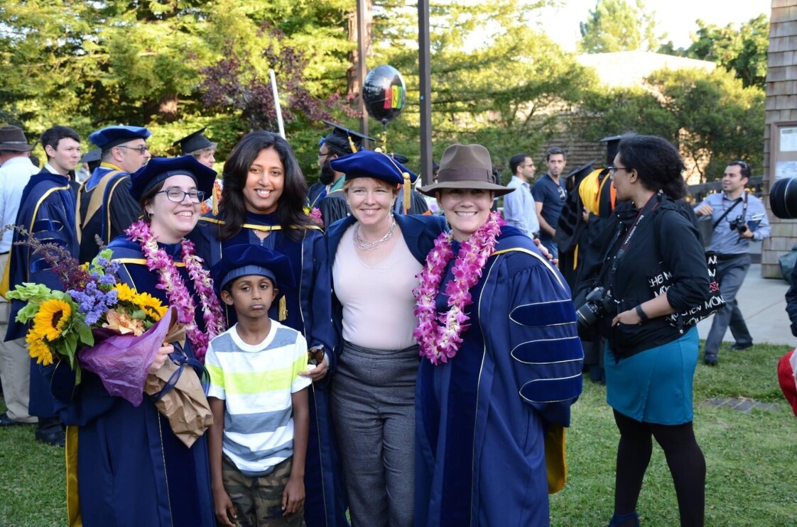 A group of college graduates in caps and gowns smile for a photo with their family members in an outdoor setting.