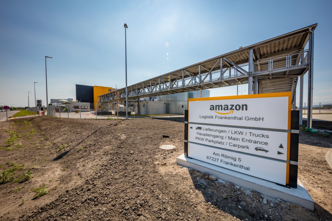Eine Außenaufnahme zeigt die Beschilderung für das neue Logistikzentrum. Pfeile auf dem Schild weisen den Besuchern den Weg zum Eingang, Parkplatz oder für die Anlieferungen. Das Schild steht auf einer frisch angelegten Außenanlage. Erde ist zu sehen. Im Hintergrund erkennt man das Außengebäude des Logistikzentrums.
