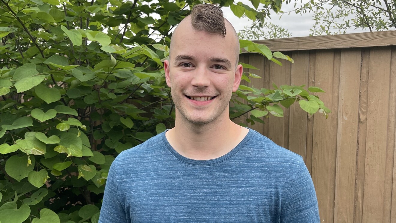 Bryce smiling, wears a blue T-Shirt and has a mohawk. He is standing in front of a bush and a tan fence.