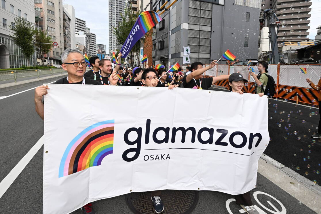 メガネをかけている男性2人と帽子を被っている女性がGlamazon Osakaの横断幕を持って、グループの先頭を歩いている。彼らの後ろには数人の人々が続いている。そのグループの右側にはたくさんのシャボン玉が飛んでいる。