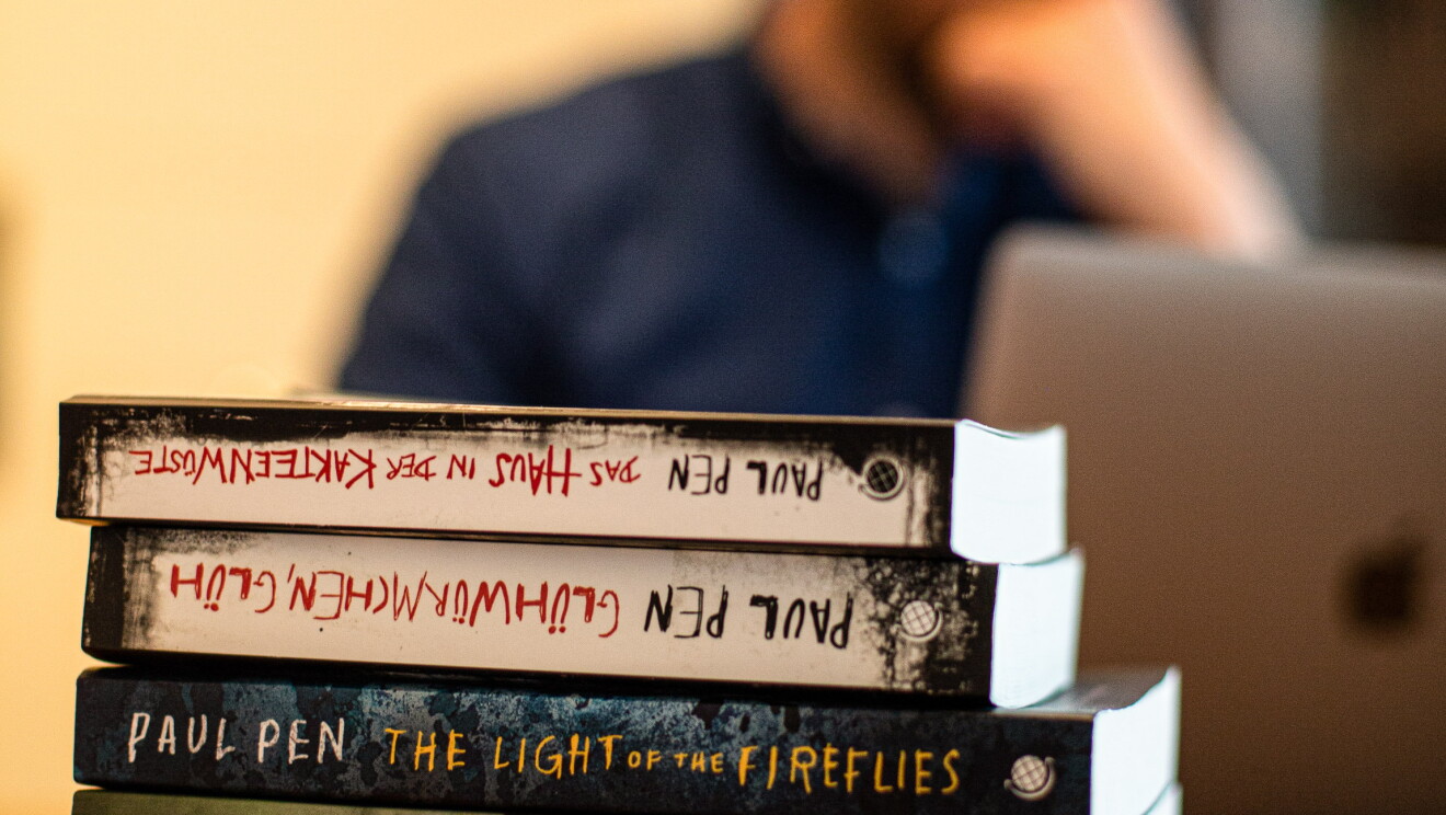 Paul Pen Paul empezó su relación con Amazon Crossing, la editorial especializada en traducciones, en 2015. Tres de sus cuatro novelas se han publicado al inglés y al alemán. En primer plano aparecen sus tres novelas traducidas al inglés y otra en alemán. De fondo y desenfocado, Paul trabajando en su ordenador. 