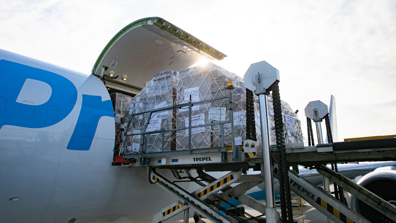 En la pista de un aeropuerto un avión con ayuda humanitaria. El avión es de color blanco con las letras en azul Prime Aire. El cielo está nublado y es el atardecer. En primer plano una grúa que carga un palé de ayuda humanitaria. El palé es de color marrón, cubierto de plástico y unas cuerdas que fijan las cajas. 