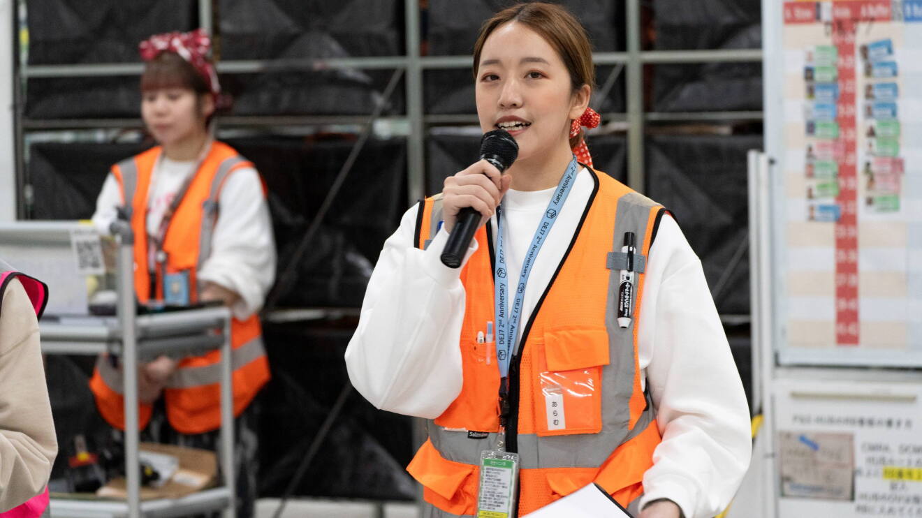 オレンジ色の作業用のベストを着ている女性がマイクで話している。ナム・アラムさん