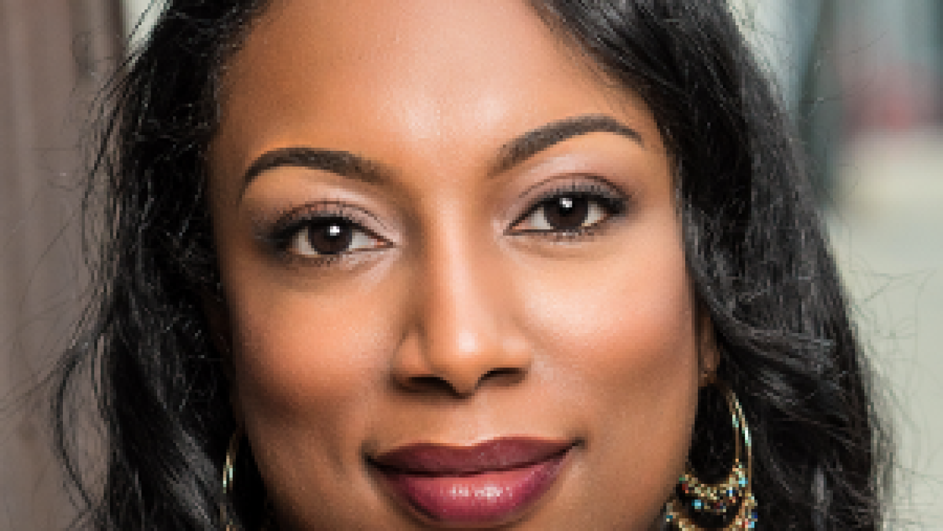 A woman with dark skin and black hair looks at the camera with a slight smile on her face