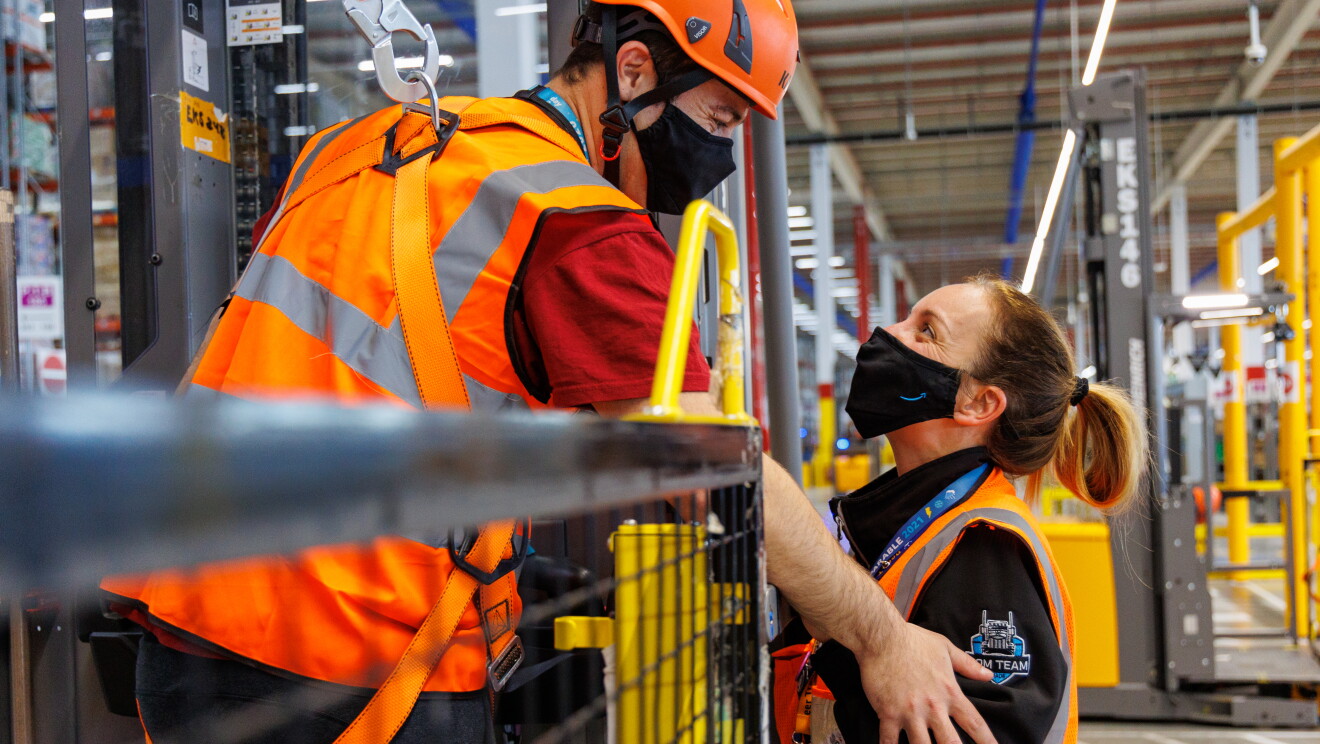 Sira Valeriano y Cristian Gamella. Foto de medio plano mirándose con mascarilla de color negro con la sonrisa azul de Amazon Es una foto interior, dentro del centro logístico de illescas (Toledo). Los dos protagonistas van con una camiseta negra, él de manga corta y ella una sudadera y él va con el pelo corto y ella largo recogido con una coleta. Él está dentro de un toro enganchado a un arnés y con su mano abraza el brazo de Sira. 