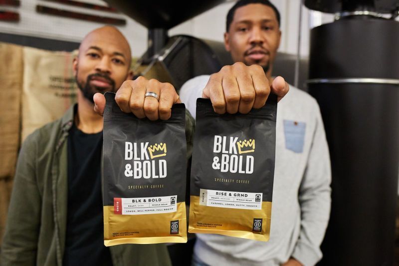 The founders of BLK & Bold hold up bags of their coffee standing side-by-side.