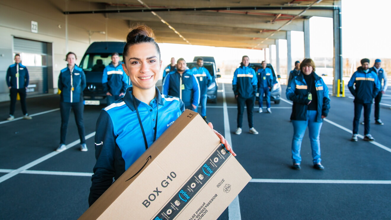 Eine zierliche junge Frau steht im Vordergrund mit einem Amazon Paket, hinter ihr sind 30 Mitarbeiter:innen verteilt auf einem Parkplatz zu sehen. Alle tragen Arbeitskleidung im Amazon Fahrer:innen Branding.