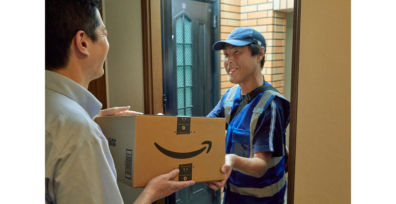 玄関口で、対応した人物にダンボール箱を手渡す配送スタッフ。箱の表面にはAmazonロゴ。配送スタッフはキャップをかぶり、ベストを着用