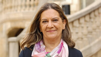 La escritora Mercedes Pinto con una chaqueta azul y un pañuelo a tonos rosa y morado. De ojos azules y pelo claro, mira a cámara sonriendo. El fondo de la foto es la escalinata del Palau Robert de Barcelona. 