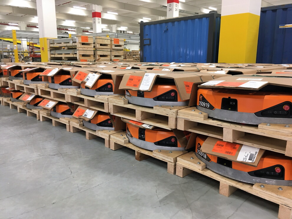 Auf Paletten stapeln sich orange-farbene Amazon Transportroboter. Sie ähneln haushaltsüblichen Saugroboter, sind aber deutlich größer.