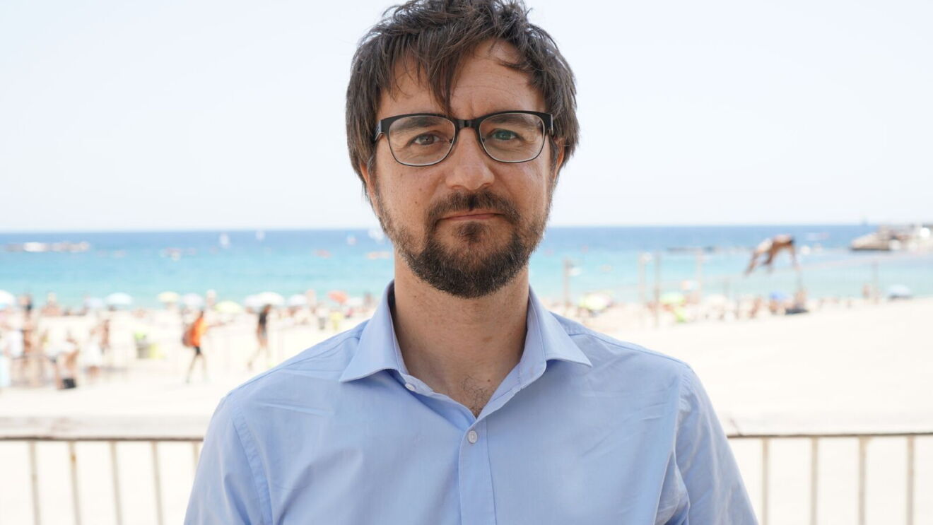 Primer plano del ingeniero del Amazon Research and Development Center de Barcelona, Hugo Zaragoza. Lleva una camisa azul, barba y unas gafas de pasta. El fondo difuminado es el de la playa de la Barceloneta. 