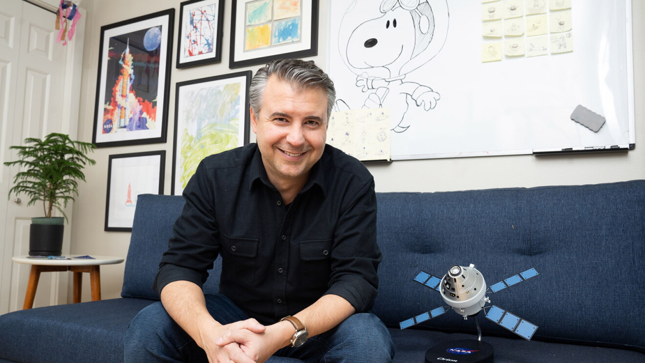 An image of a man smiling for a photo while sitting on a sofa in his office. Next to him is a small satellite device.