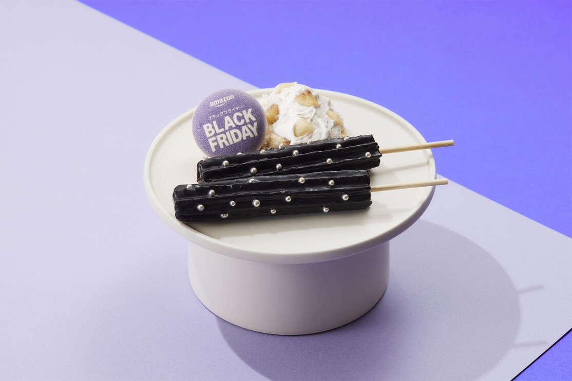 高台が付いた白い器に串にささった黒いチュロスが2本置かれている。チュロスの表面には銀色のアラザンが点在している。その後ろにホイップクリームとアーモンドスライス。Black Fridayと書かれたクッキー