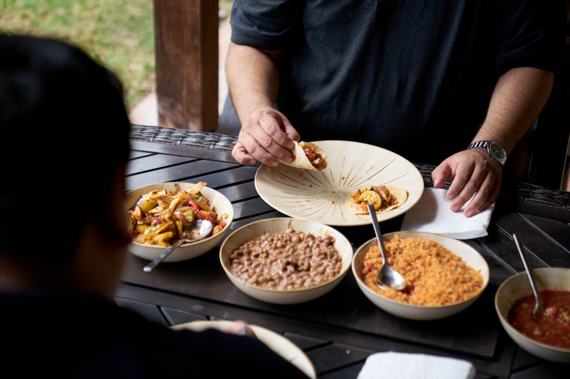 Two men sit at a table and eat tacos.