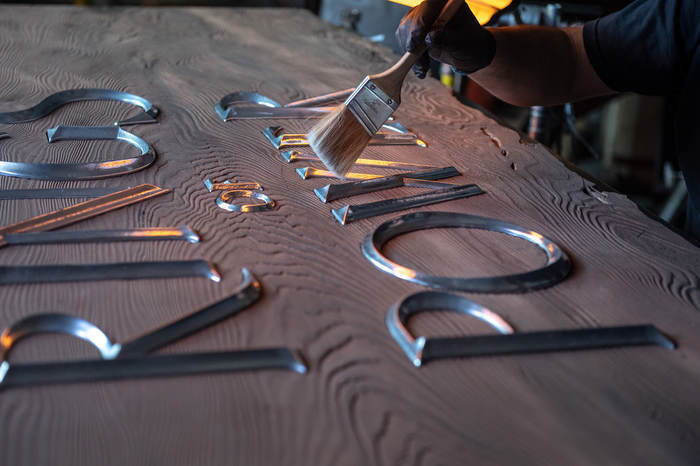 An image of a wooden plank with metal letters on it that read "The Lord of the Rings: Rings of Power." There is a paint brush brushing off the letters. 