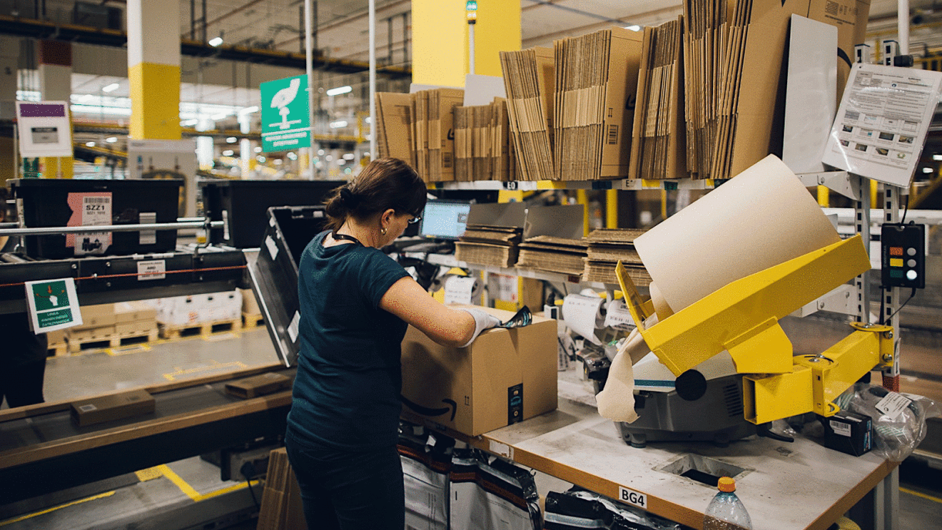 A woman stands at a desk full of boxes and tape. She is boxing up an item to be shipped.