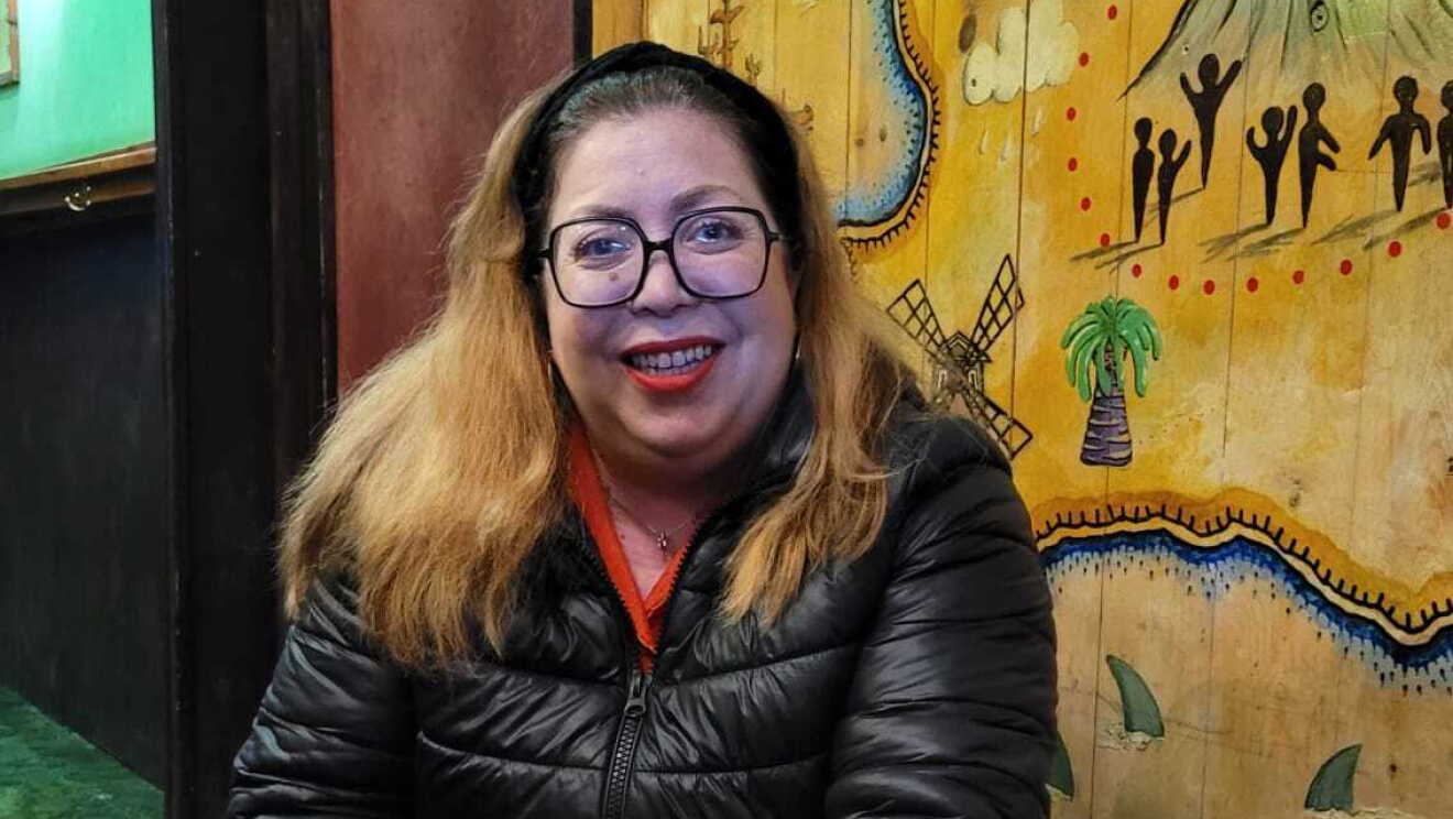 Anna Cánovas con su perro Yonce en la calle. El perro es un labrador de color marrón muy claro. Ella está agachada al lado del perro. Lleva el pelo largo hasta después de los hombros. El pelo de decolorado. Lleva gafas de pasta negras. Una chaqueta plumón de color negro, debajo un jersey de color rojo, unos pantalones negros y una deportivas básicas. De fondo una pared de madera con dibujos étnicos, como pintados a mano. 