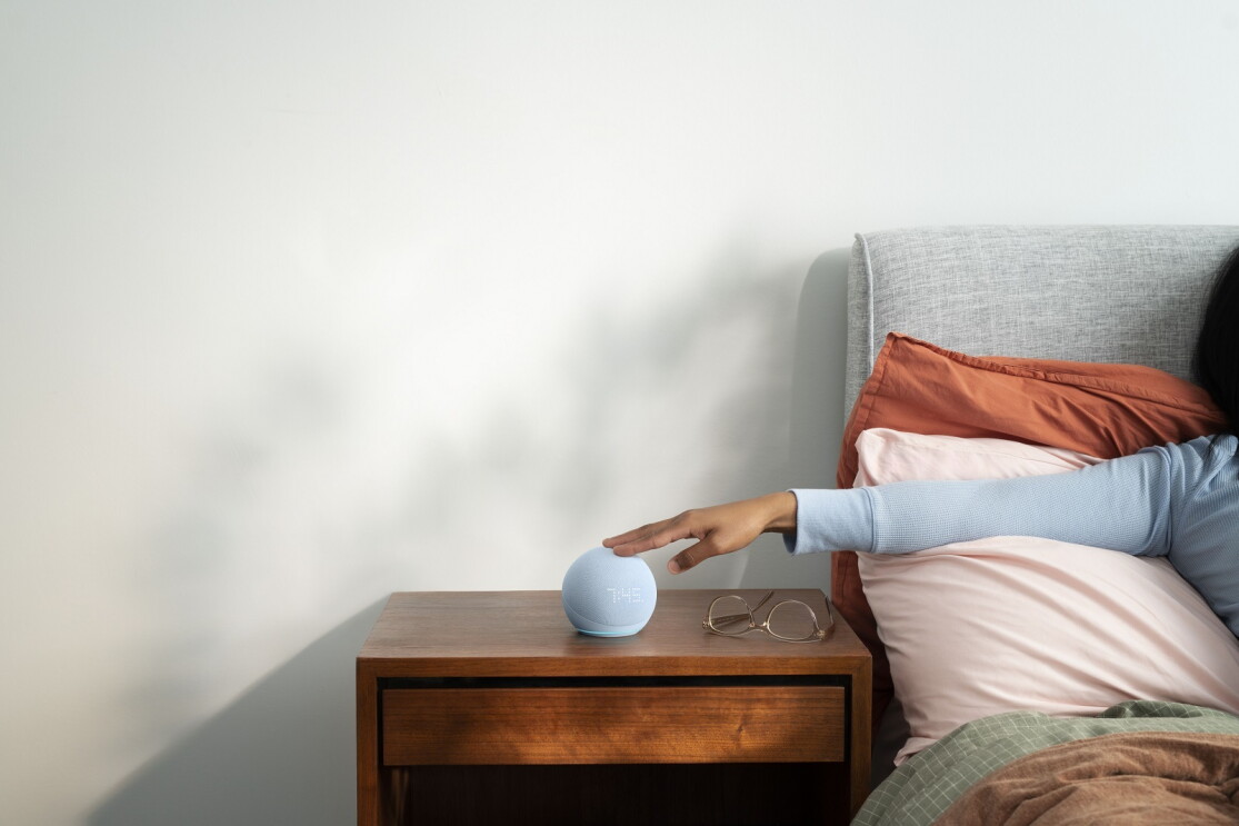Ein blauer Amazon Echo Dot steht auf einem Nachttisch neben einem Bett. Aus dem Bett sieht man einen Arm, der auf das Gerät tippt.