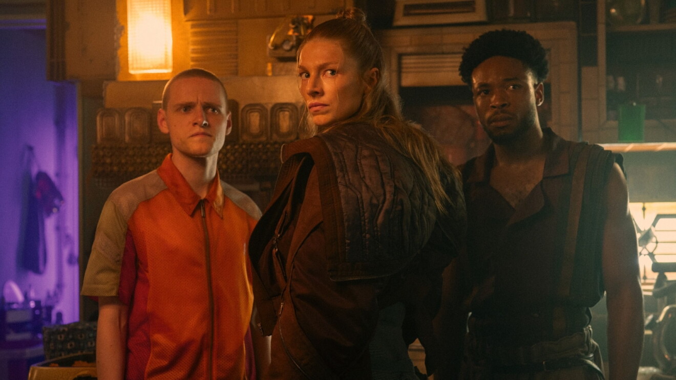 Image from 'Blade Runner 2099' of three characters stand together in a dimly lit, industrial interior with purple and amber lighting