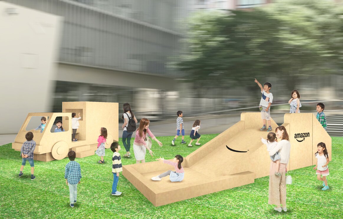 芝生広場に設置された段ボール遊具。子どもが乗って遊べる配送トラック型の遊具と滑り台の2つ。遊具はベージュ色でAmazonのロゴが描かれている。遊ぶ子どもたちと見守る保護者がいる。