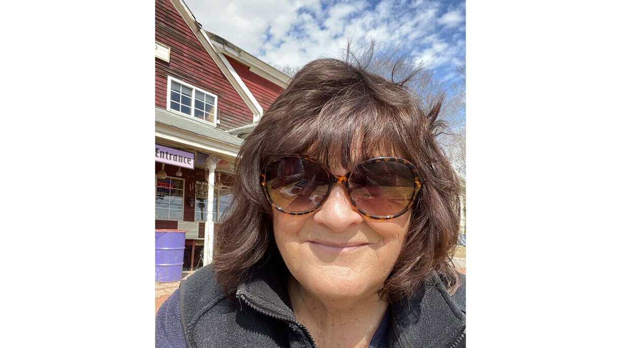 A selfie image of Kate. She is standing in front of a red and white wooden building wearing sunglasses.