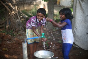 An image of two children standing at a water spigot. They are smiling and holding their arms out to cup the water in their hands.  