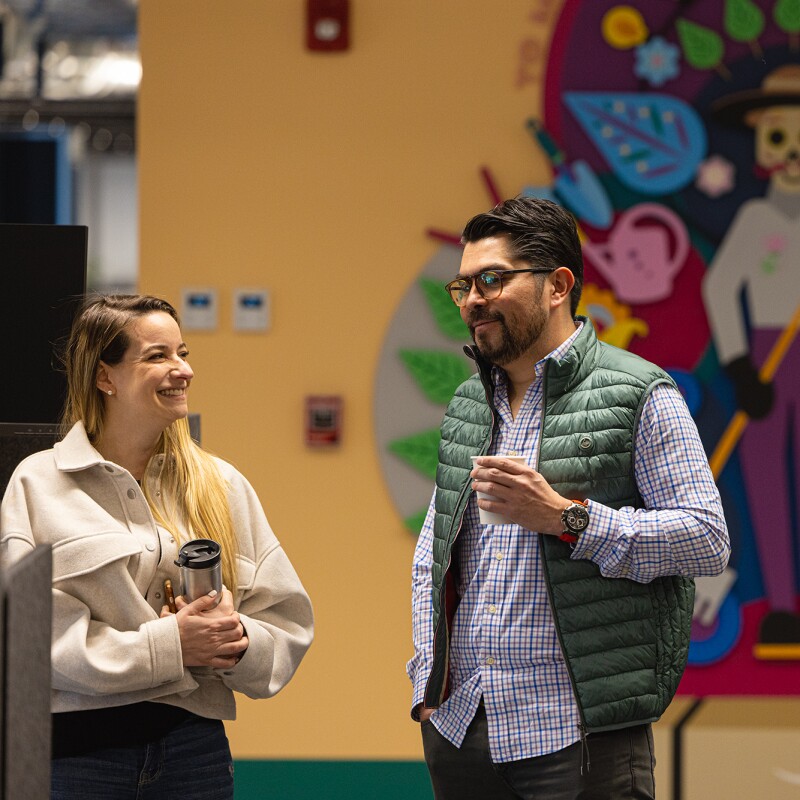 De Una foto tomada en las oficinas de Amazon. De fondo un dibujo estampado en la pared que parece del Día de los Muertos. EN la parte derecha las mesas con las sillas. En la parte izquierda hay dos personas hablando de pie. Una mujer rubia con el pelo largo y agarrando un recipiente de mate. Lleva una chaqueta de color blanco y un pantalón marrón. Él va con una camisa de cuadros , un chaleco verde, barba y perilla oscura y gafas. Está agarrando un vaso de café.