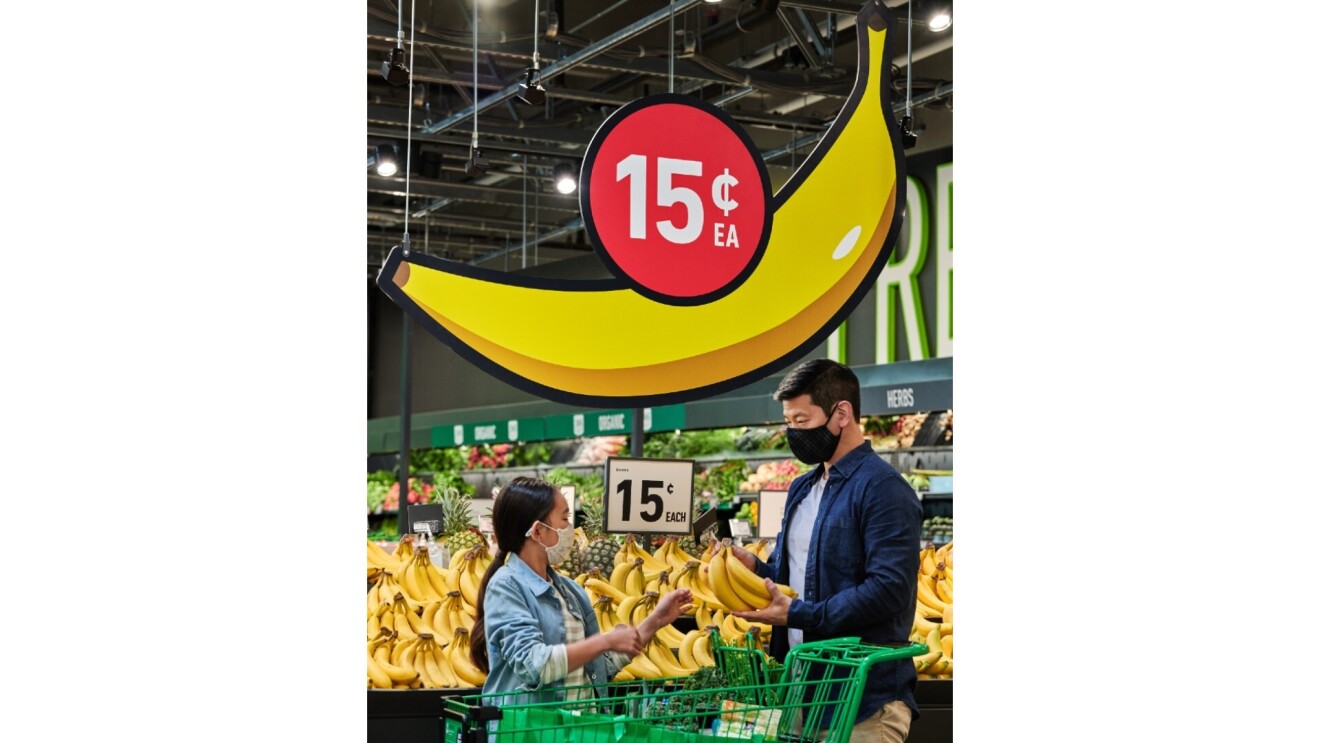 A father and his daughter shop for bananas at Amazon Fresh. Above them is a sign in the shape of  a banana with 15 cents above it.