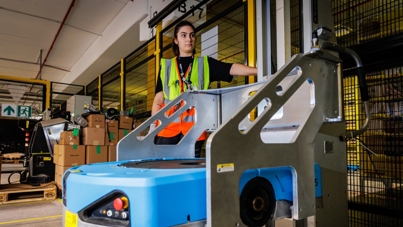 Dentro de un centro logístico, está una mujer con un elevador para reparar un robot de color azul. Está de pie, lleva el pelo recogido con un moñparrodillada mirando un robot está colocado en una mesa. Ella tiene el pelo oscuro largo y recogido en una cola. Van con un camiseta negra de manga corta y un chaleco amarillo.