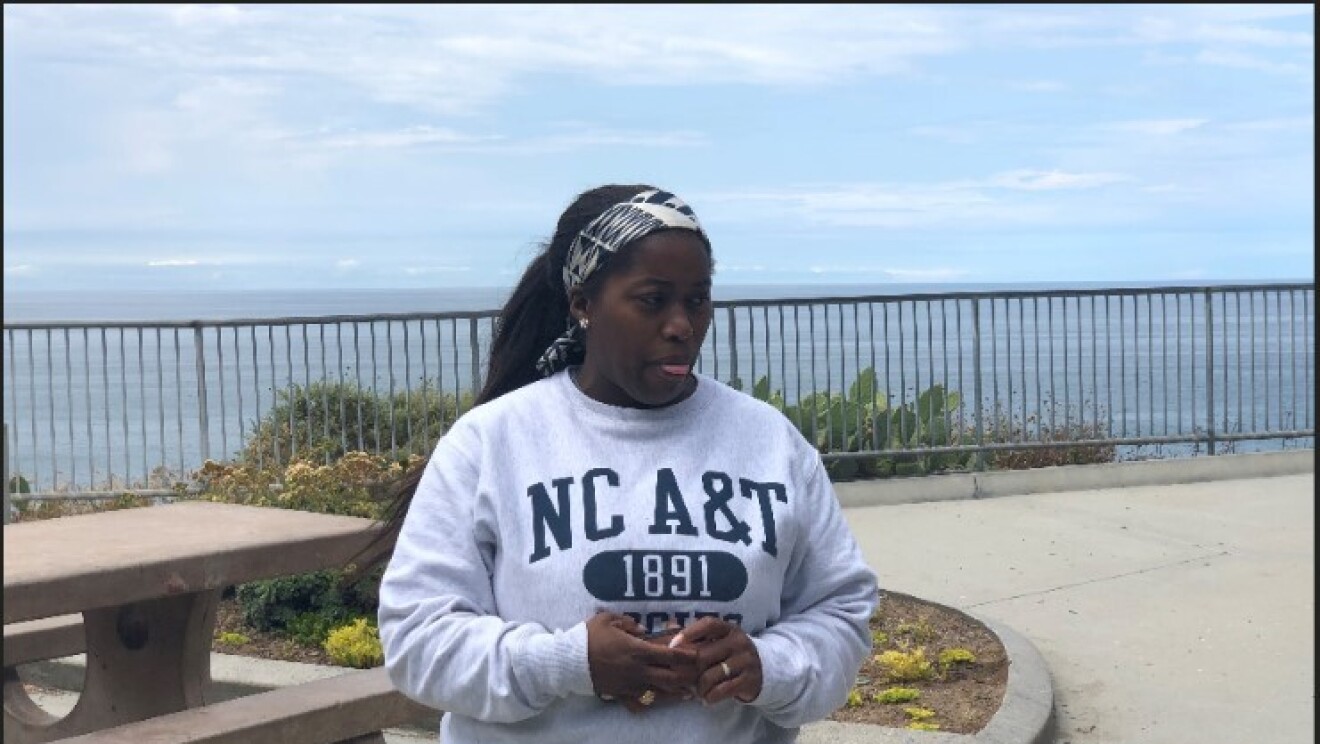 A woman wearing a sweatshirt stands on a boardwalk near a large body of water.