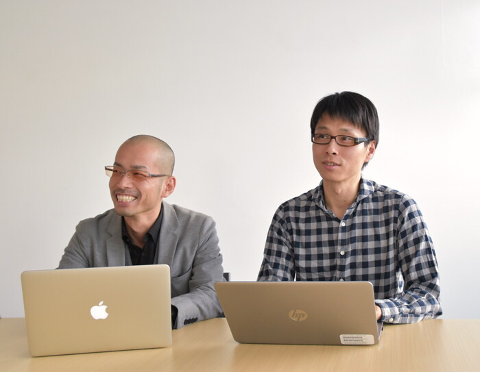 白い壁を背に、ベージュ木目のデスクに並んで向かう男性2人。共に銀色のノートパソコンを開いている。