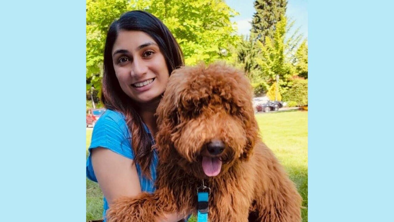 Poonam smiling and sitting on a blanket in a park while holding her mini goldendoodle, Clark. 