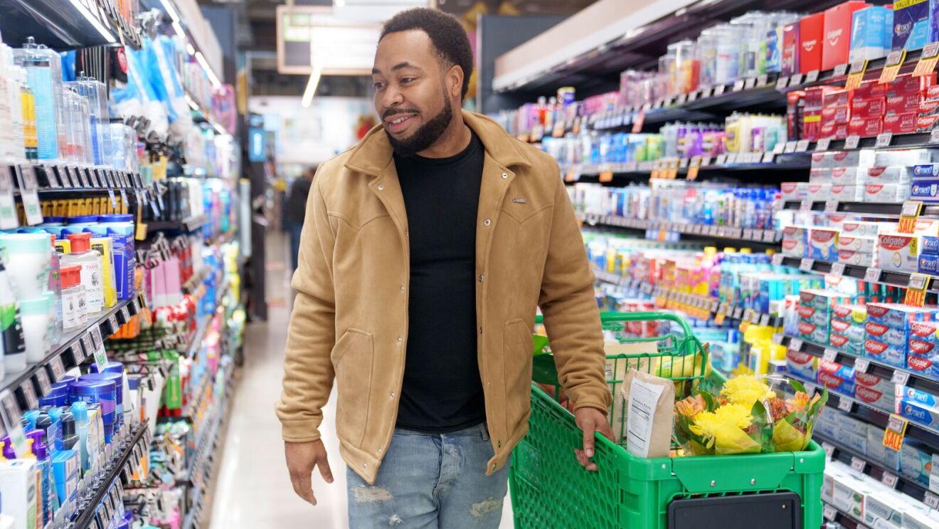 Smiling shopper in tan jacket pushing green cart down grocery aisle