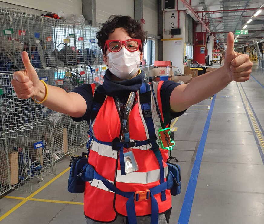 Mezzo busto di una dipendente Amazon in un magazzino. Ha le braccia alzate e con entrambe le mani fa il segno dell'OK con i pollici in alto. Ha i capelli corti e indossa un paio di occhiali da vista rossi. 