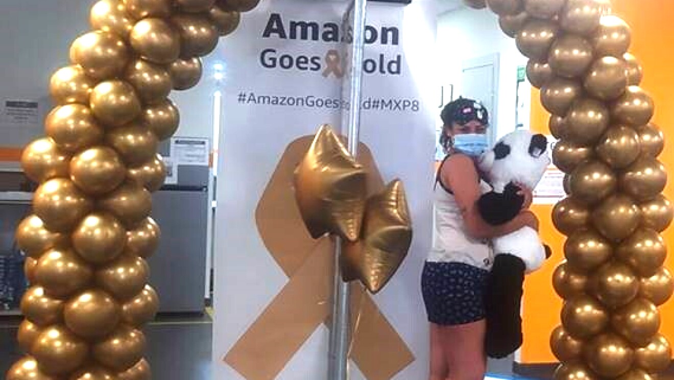 Dipendente Amazon posa con un peluche a forma di panda sotto un arco di palloncini dorati e accanto un cartellone con la scritta Amazon Goes Gold 2021