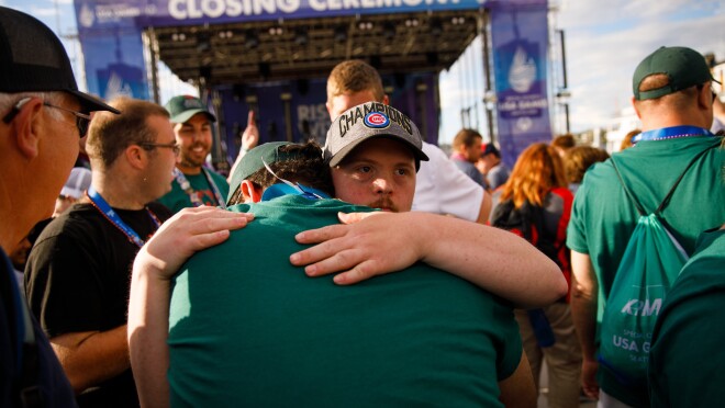 一个男人拥抱另一个男性,庆祝美国奥运会闭幕式。在后台,更多活动的参加者出现庆祝,舞台设置在远处。