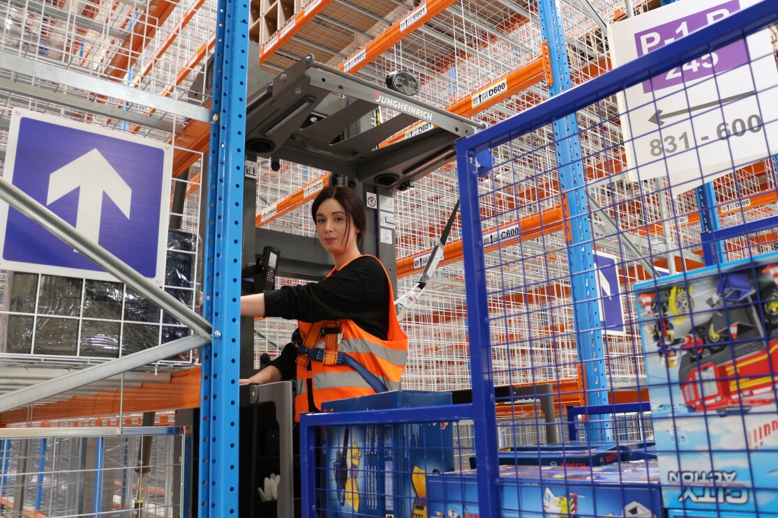 Ein Logistikmitarbeiterin fährt einen Vertikal-Kommissionieren. Im Hintergrund sieht man die neuen Hochregale.  
