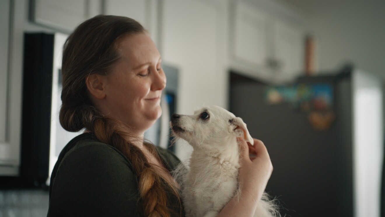 An image of a woman holding a small, white dog. She is looking down at the dog and smiling. 