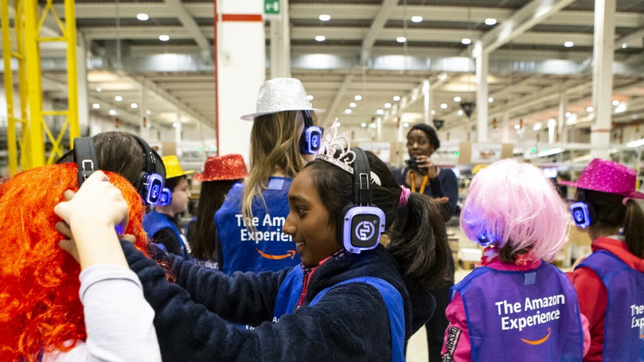 Un gruppo di bambini in visita in un centro di distribuzione ascolta la tour leader che illustra loro le caratteristiche del centro. I bambini indossano parrucche e cappelli colorati per carnevale, insieme alle cuffie per ascoltare le spiegazioni del tour. Sono tutti rivolti di spalle rispetto alla camera, tranne una bambina in primo piano, girata a sinistra, che gioca con la cuffie della bambina accanto a lei. 