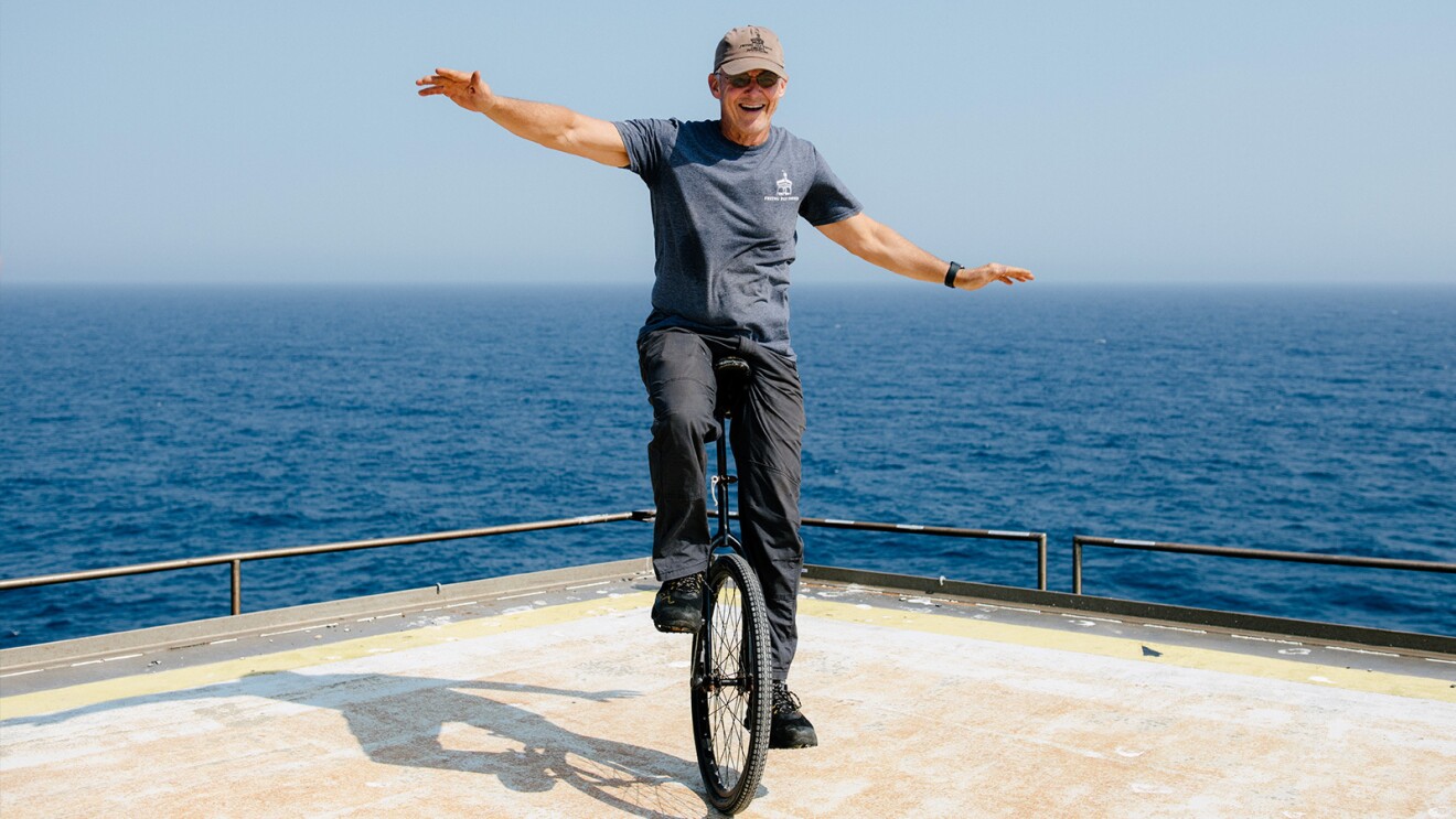 A man smiles as he works to stay balanced on a unicycle with the ocean behind him.