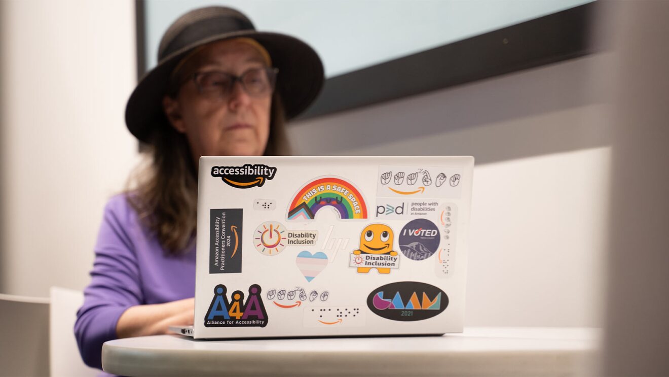 Laptop covered in accessibility and disability inclusion stickers, foreground of person in purple shirt and hat