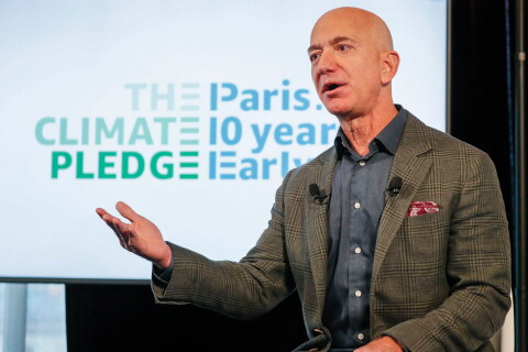 De fondo el cartel the The Climate Pledge, Paris 10 years early. Jeff Bezos en primer plano con una mano levantada. Una amerciana verde y gris a cuadros y una camisa azul. En la americana lleva un pañuelo rojo.  
