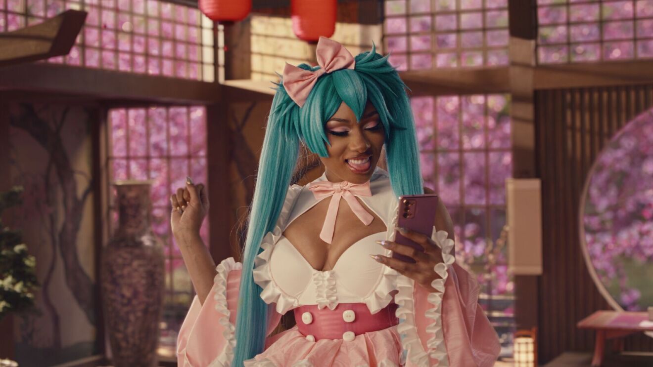 An image of Megan Thee Stallion in a cosplay outfit while smiling and looking at her phone. She is wearing a bright blue wig with long pigtails and an Asian-inspired outfit while standing inside a dojo.