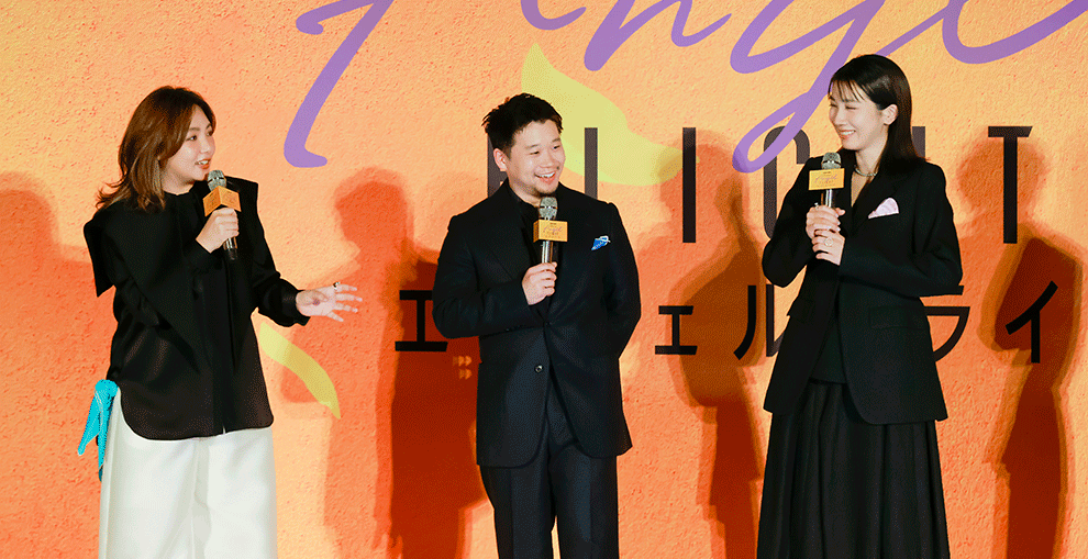 オレンジ色の壁の前、オレンジのステージの上に、黒い服を着て横一列に並んだ3人の人物。全員マイクを手に持って会話をしている。