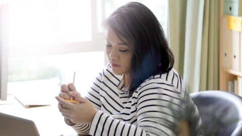 A woman writes note on a sticky notepad with a pen. Behind her, light steams through a window. 