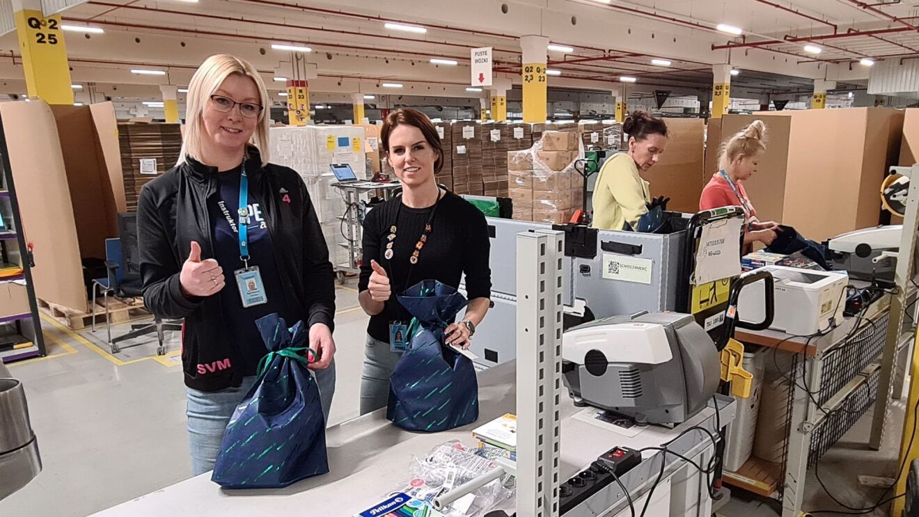 Zwei Mitarbeiterinnen von Amazon mit blauen Taschen in einem Logistikzentrum für eine Spenden-Aktion.