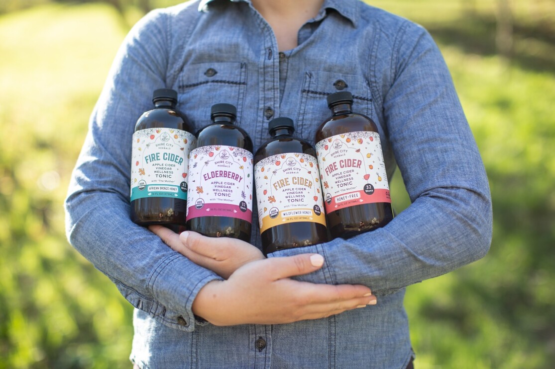 A person holds four bottles of Fire Cider in their arms while standing outside.