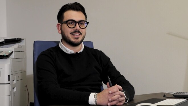 Mezzo busto di Mario Laddaga, seduto ad una scrivania con una penna tra le mani. Alle sue spalle, a sinistra, una fotocopiatrice